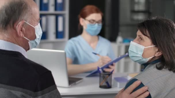 Äldre par i medicinska masker i ansiktet i samråd med kvinnlig läkare under virus och infektion diskuterar hälsotillstånd medan sitter på sjukhus — Stockvideo