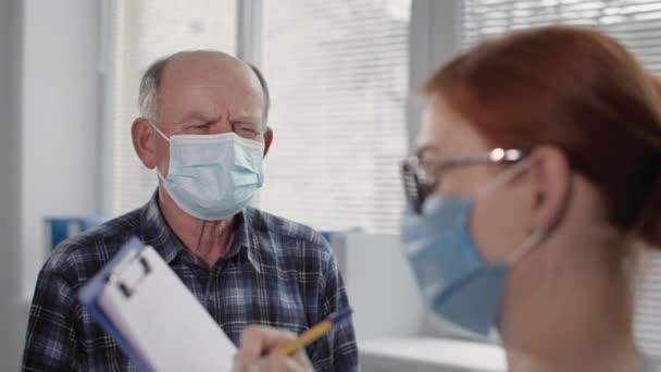 Alter männlicher Patient mit medizinischer Maske, um sich vor dem Virus der Infektion zu schützen, an der Rezeption einer Ärztin diskutiert die Behandlung — Stockvideo