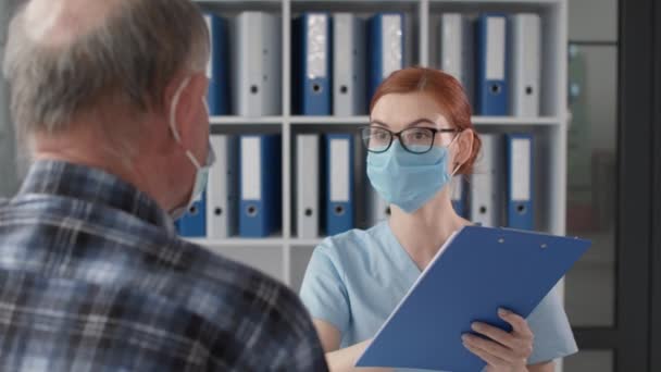 Vrouwelijke arts met medisch masker en bril praat met een oudere man over zijn gezondheidstoestand tijdens het onderzoek van de patiënt in het ziekenhuis kantoor — Stockvideo
