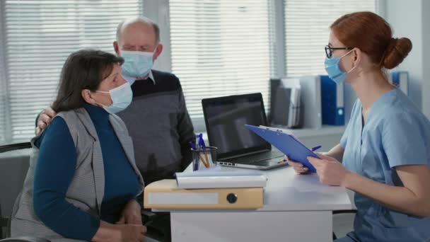 Jovem médica vestindo uma máscara médica para proteger contra o coronavírus discute a saúde de pacientes idosos durante exame médico no consultório hospitalar — Vídeo de Stock