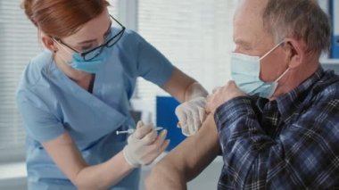 Maske ve eldiven takan profesyonel hemşire, yaşlı bir adama salgın sırasında virüs ve enfeksiyondan korunmak için aşı enjekte ediyor.