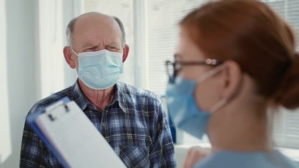 Kadın doktorla resepsiyonda yüzüne tıbbi maske takan yaşlı bir adam klinikte tedaviyi tartışıyor. — Stok video