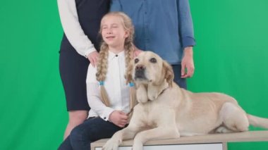 Labrador köpeği ve yeşil krom arka planda anne ve babasının gülümseyen ve kameraya bakan gülümseyen kız çocuğunun portresi.