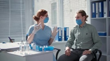 Tıbbi maske ve eldiven giyen hemşire, hastane ofisindeki genç adama, tehlikeli bir virüse karşı modern bir aşı gösteriyor.