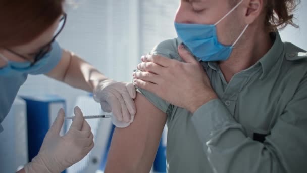 Vrouwelijke arts vaccineert jonge gemaskerde man met spuit en naald tijdens een coronavirus pandemie — Stockvideo