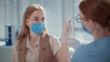Doktor, tıbbi maske takan genç bayan hastaya ilaç iğnesi yapmak için şırınga ve şırınga kullanıyor.