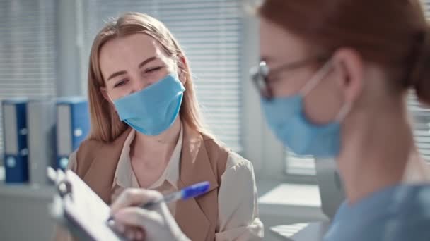 Γυναίκα ασθενής που φοράει ιατρική μάσκα για την προστασία από τον ιό και τη μόλυνση υποβάλλεται σε εξέταση και μετρά τη θερμοκρασία με θερμόμετρο χωρίς επαφή κατά τη διάρκεια του διορισμού των ιατρών στο νοσοκομειακό γραφείο — Αρχείο Βίντεο