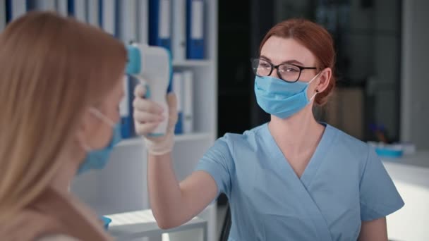 Medico che indossa maschera e guanti esamina paziente femminile misura la temperatura con termometro senza contatto — Video Stock