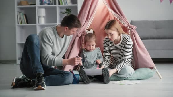 Desarrollo del niño, los padres cariñosos junto con su hija amada se dedican al desarrollo del niño y dibujan con lápices de colores en el fondo de papel de bigwam y miran a la cámara — Vídeos de Stock