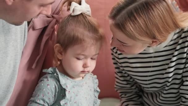 Kindheit: Junge männliche und weibliche Eltern küssen ihre kleine Tochter liebevoll und zärtlich, wenn die Familie zu Hause sitzt — Stockvideo