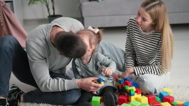 Padres de familia amorosos jóvenes con hija disfrutan de vacaciones conjuntas besando a su hijo femenino y jugando con constructores de plástico de colores en la habitación de los niños sentados en el suelo — Vídeos de Stock