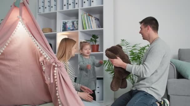 Parenting, happy husband and wife have fun with their little cheerful daughter with a soft toy and kiss each other while sitting in the childrens room with a wigwam — Stock Video