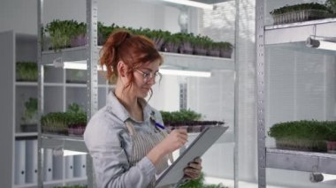 Genç iş kadını konteynırlarda mikro yeşillikler yetiştiriyor ve rafların arka planında dururken filizlerin durumunu inceliyor ve kaydediyor, modern sera
