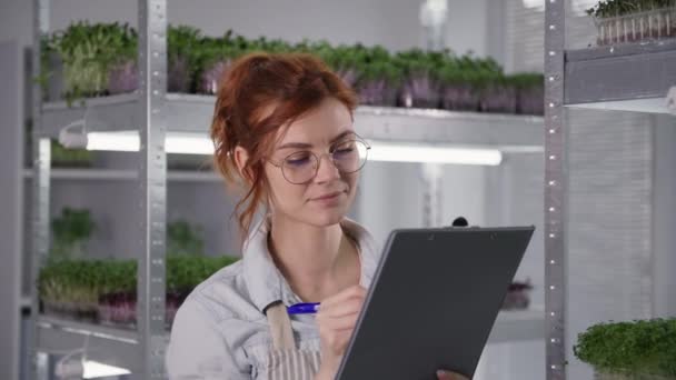 Organiczny biznes, słodkie młoda kobieta rośnie mikro zielony w pojemniku na półkach w nowoczesnej szklarni sprawdza stan kiełków i robi notatki — Wideo stockowe