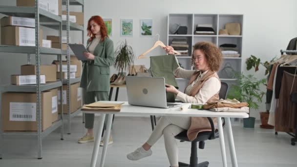 Онлайн-торгівля, молода жінка продає одяг клієнту за допомогою ноутбука і показує штани на вішалці з жінкою, яка готує доставку на задньому плані в магазині одягу — стокове відео