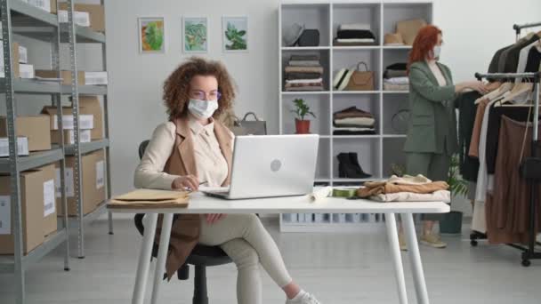 Work during pandemic, female seller wearing medical mask communicates with buyer via video link and shows product online in clothing store during pandemic — Stock Video