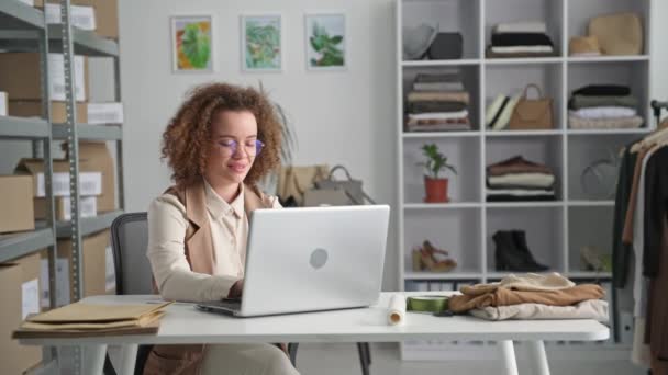 Pequeno negócio, retrato de uma jovem mulher tomando ordens on-line em um laptop enquanto se senta em uma mesa em uma loja de roupas — Vídeo de Stock