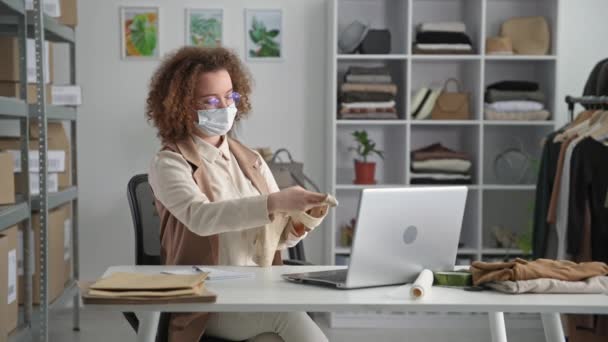 Trabajo en la imagen, joven emprendedora con máscara protectora toma una orden en una tienda en línea utiliza el ordenador portátil mientras trabaja en cuarentena, sonríe y mira a la cámara — Vídeo de stock