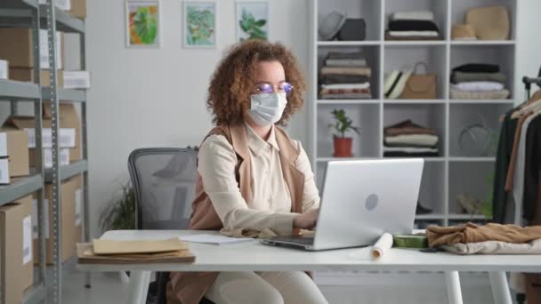 Tıp maskeli başarılı bir satıcı online bir mağaza aracılığıyla iyi satışlar yapıyor ve laptopla masada otururken ellerini kaldırıp karantinada çalışıyor. — Stok video