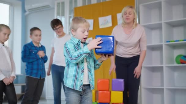 Garçon avec le syndrome du duvet joue à des jeux éducatifs avec les enfants et en passant blocs colorés les uns aux autres dans une salle de classe inclusive — Video