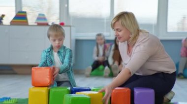 Bayan öğretmen Down sendromlu çocuklar için gelişim faaliyetleri yürütüyor ve engelli çocukların kapsamlı bir okulda ders sırasında renkli tuğlalar eklemelerine yardımcı oluyor.