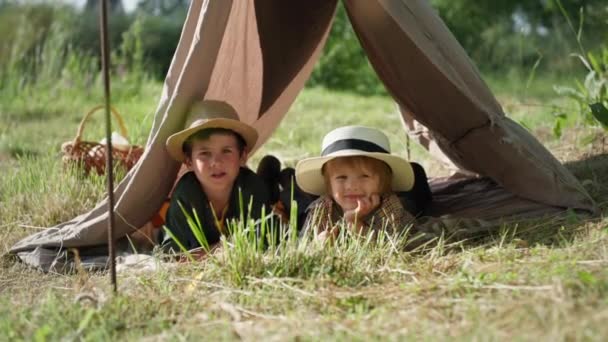 Retrato de lindos niños pequeños en el sombrero se encuentran en el césped verde en un wigwam mientras descansa en el campo en las vacaciones de verano, mirando a la cámara — Vídeos de Stock