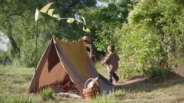 Juegos activos para los niños, hermanos felices se divierten corriendo alrededor de la tienda de campaña en el césped verde durante sus vacaciones de verano fuera de la ciudad — Vídeos de Stock