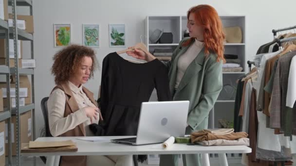 Online försäljning, ung flicka entreprenör tillsammans med en kollega visa kläder till köpare genom videosamtal på bärbar dator när du sitter vid bordet i fashionabla butik — Stockvideo