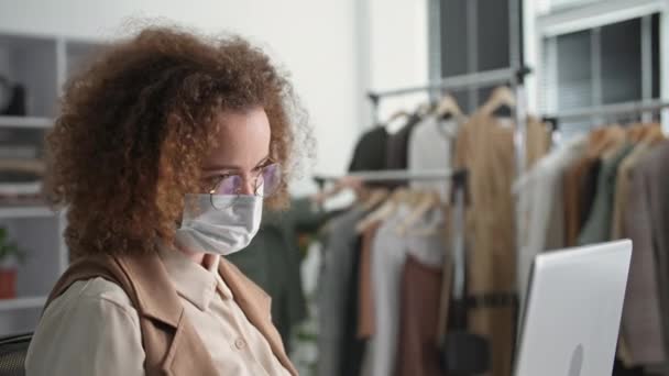 Joven mujer creativa en una máscara médica trabaja en una tienda de Internet y se comunica con un cliente a través de un enlace de vídeo que vende bienes utilizando tecnologías modernas, el trabajo durante la cuarentena — Vídeo de stock