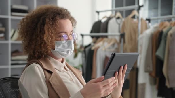 Portret van een jonge vrouw in een medisch masker en bril gebruikt een tablet voor online verkoop in een internetwinkel terwijl ze in een modeboetiek zit — Stockvideo
