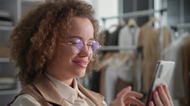 Gülümseyen, gözlüklü genç bir kadın, bir giyim mağazasında otururken internetten mal satarken cep telefonunu dinliyor.