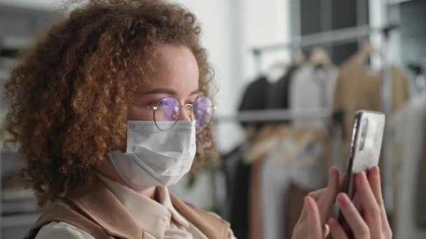 Precauciones, linda mujer joven en máscara médica para proteger contra el virus y la infección utiliza las redes sociales y grifos teléfono inteligente mientras que la venta de bienes a través de Internet mientras está sentado en la tienda de ropa — Vídeo de stock