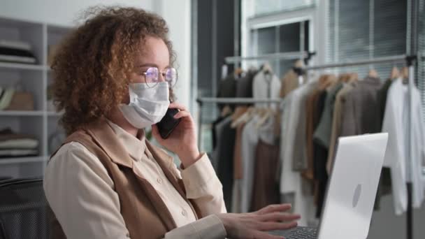 Karantinada küçük bir işletme, tıbbi maske takan başarılı genç bir iş kadını müşterisiyle akıllı telefondan konuşuyor ve salgın nedeniyle dizüstü bilgisayara sipariş veriyor. — Stok video