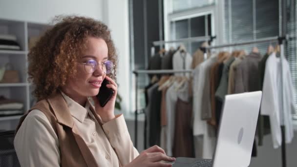 Портрет молодої жінки-продавця, що розмовляє з клієнтом на мобільному телефоні з клієнтом, приймаючи замовлення в інтернет-магазині, використовуючи ноутбук в магазині одягу — стокове відео