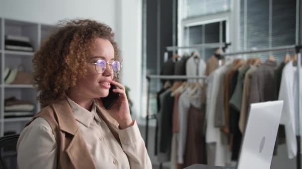 Роздрібна торгівля, красивий жіночий менеджер спілкується на смартфоні, приймаючи замовлення онлайн в інтернет-магазині на ноутбуці, сидячи в модному бутіку — стокове відео