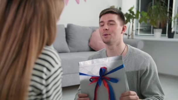 Ευτυχισμένος νεαρός άνδρας με ένα δώρο στα χέρια του ευχαριστεί την αγαπημένη του σύζυγο και την αγκαλιάζει κατά τη διάρκεια του εορτασμού της Ημέρας Πατέρων στο σπίτι — Αρχείο Βίντεο