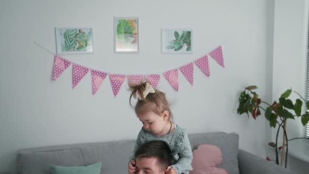 Pai e criança, um jovem brinca em um lugar com sua filha em torno de seu pescoço e senta-se durante a diversão em família em casa — Vídeo de Stock