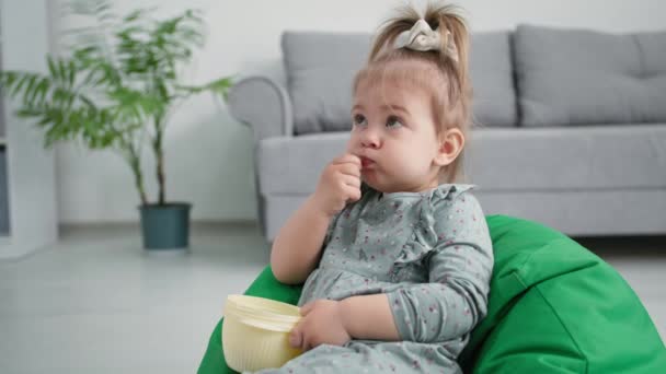Infância, bonito criança do sexo feminino com prazer come deliciosas bagas maduras enquanto faz um lanche enquanto está sentado em uma poltrona macia em casa — Vídeo de Stock