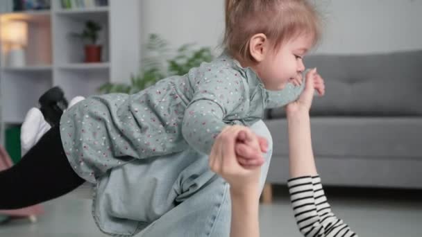 Happy little girl has fun with mom while playing the plane, joyful female parent kissing her child with pleasure while lying on floor in room — Stockvideo