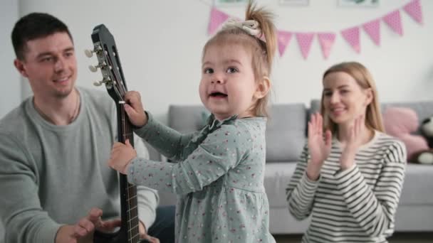 Gyermekkor, kis boldog női gyermek gitárral a kezében érezd jól magad a szülőkkel a családi szabadidő alatt otthon — Stock videók