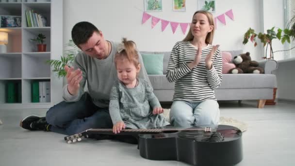 Gelukkig familie, klein mooi meisje spelen met muziekinstrumenten in achtergrond mam klapt handen en papa kust zijn vrouw terwijl ontspannen thuis — Stockvideo