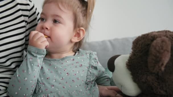 Portrait of charming little girl with pleasure eating cookies while sitting in her mothers arms in children room, close-up — Stockvideo