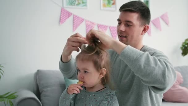 Fatherhood, loving caring male parent does his daughter hair and ties her hair up with an elastic band, close-up — Stockvideo