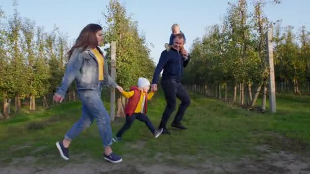 Los padres y los niños, la mujer y el hombre con un pequeño niño varón alrededor de su cuello, toman de la mano a los niños mientras corren alegremente por la carretera en el jardín en el campo — Vídeo de stock