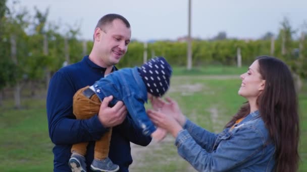 Petit bébé assis dans les bras du père et demande à sa mère, papa joyeux passé son fils à la femme tout en marchant dans le verger — Video