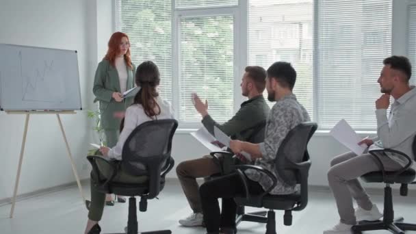 Jovem empresária faz uma apresentação de novo projeto em flipchart para mulheres e homens empregados, jovem levanta as mãos e pede perguntas durante uma reunião de negócios no escritório — Vídeo de Stock