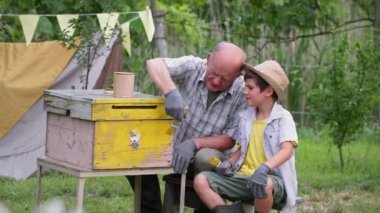 Arıcıların ailesi büyükbaba ve torun bahçedeki yeşil ağaçların arka planında oturan kovanları fırça ve sarı boyayla boyarlar.