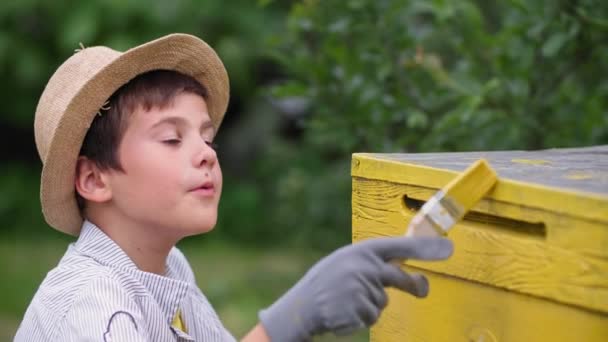 Счастливый мальчик счастливо рисует пчелиный улей желтой краской, помогая своему деду-пчеловоду во время отдыха в деревне — стоковое видео