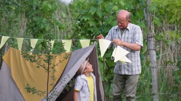 Loisirs de plein air, enfant mâle joyeux en chapeau de paille et son grand-père décorer wigwam avec des drapeaux pendant les canules dans la campagne toile de fond d'arbres verts — Video