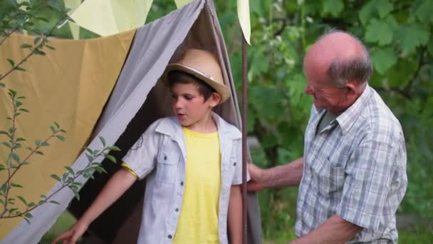 Crianza, cuidado abuelo se divierte con lindo niño varón hablando cerca wigwam contra árboles verdes fondo del jardín — Vídeo de stock